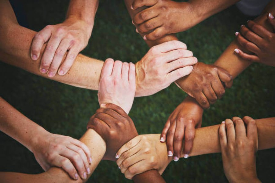 Looking down at many mixed hands, each clasping the wrist of another person and forming an interlinked human chain or mesh.